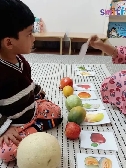 A teacher holds up a picture card for a child to match with a real fruit. This simple game is an effective way to build vocabulary and object recognition.