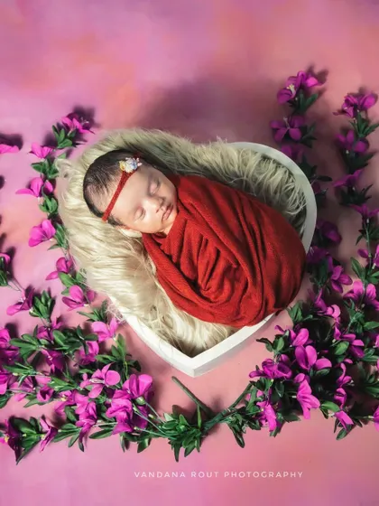 A full view of the newborn wrapped in red, nestled among purple flowers. This artistic composition is a beautiful example of a posed newborn studio session.