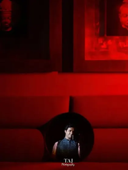 A creative framing shot, where the groom is seen through a circular opening in a vibrant red couch. This kind of composition is playful, artistic, and creates a visually interesting portrait.