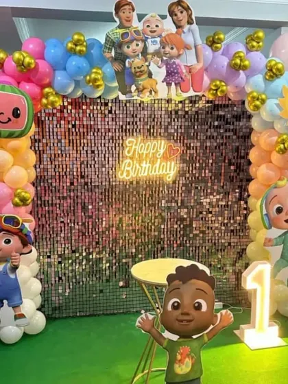 A bright and cheerful Cocomelon party with a shimmer wall. This setup features a colorful balloon arch, several character cutouts, a neon sign, and a light-up number '1'.