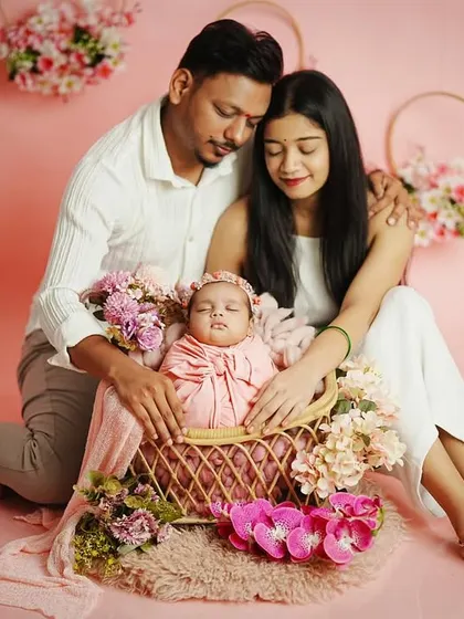 A serene family portrait with the parents and their sleeping newborn. The coordinated white outfits and pink floral theme create a harmonious and gentle image.