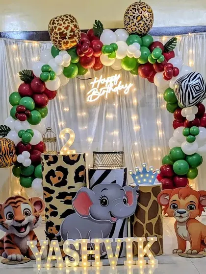 A cute jungle theme for a second birthday. This setup includes an arch with animal print balloons and cutouts of a giraffe, tiger, elephant, and lion.