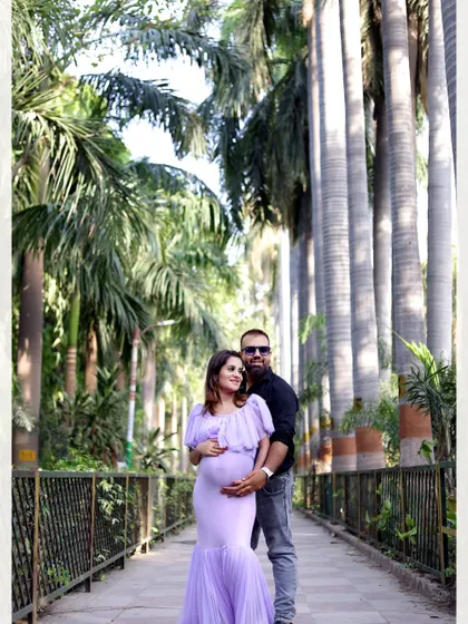 A full-length couple's portrait on a walkway surrounded by tall palm trees. The mother-to-be wears a lovely lavender mermaid-style gown.