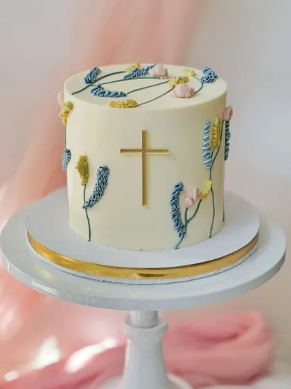 A tasteful cake for a religious ceremony, such as a baptism or first communion. It features delicate piped wildflowers and a simple, elegant gold cross.