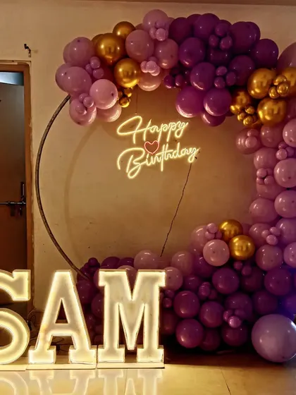 A personalized birthday decoration with large light-up letters spelling "SAM" and a purple and gold balloon ring.