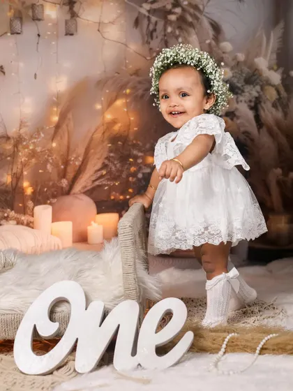 A joyful first birthday portrait for Alora. Her happy smile is the centerpiece of this photo, complemented by a rustic, warm-toned backdrop and a delicate floral crown.