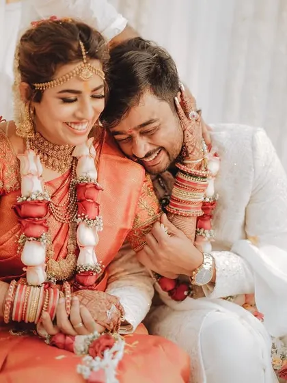 A tender, candid moment during a wedding ceremony. The groom rests his head on his bride's shoulder, both of them sharing a happy, relaxed laugh.