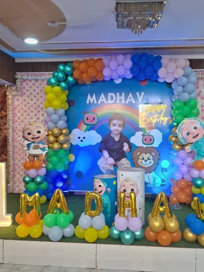 A vibrant Cocomelon themed birthday setup for Madhav's first birthday. The decoration features a colorful balloon arch, a personalized backdrop, and balloon letters spelling out the name.
