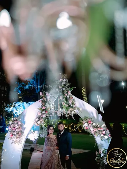 An artistic, through-the-glass shot of a couple standing under a modern white arch decorated with flowers.