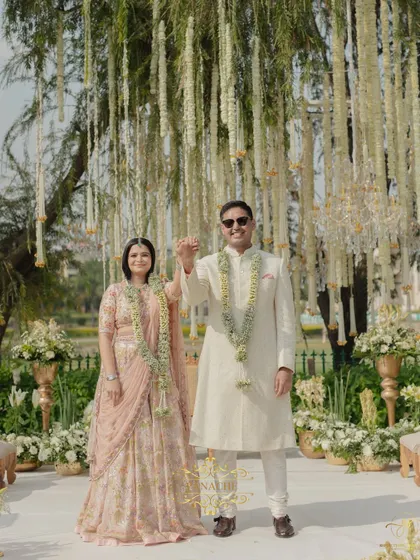 The couple raises their hands in a gesture of celebration after their Jaimala. The backdrop of hanging white florals from the trees creates a stunning, ethereal effect.