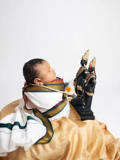 A color version of the devotional pose, with the baby wrapped in a traditional white dhoti with a green and gold border, looking towards the idols.