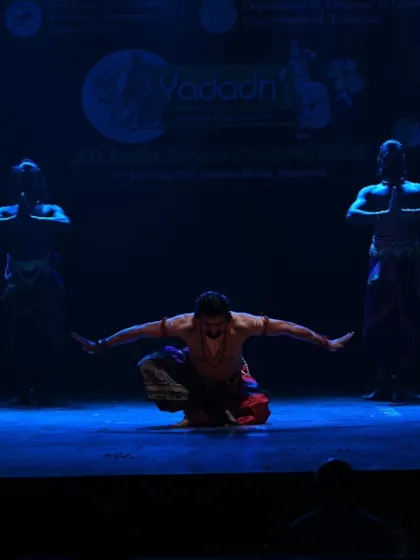 A dramatic low-crouch pose under stark blue lighting. This moment in the performance emphasizes the control and muscular tension inherent in the Chhau dance style.