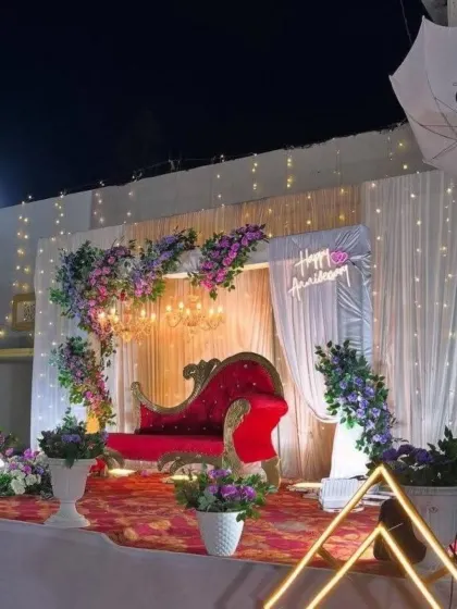 A beautiful anniversary stage at night, featuring a red velvet chaise lounge, purple and white floral decor, and warm fairy lights for a magical glow.