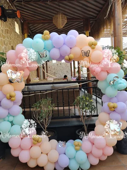 A circular balloon arch in pastel rainbow colors, adorned with silver butterfly accents, creates a beautiful photo opportunity for an outdoor event.