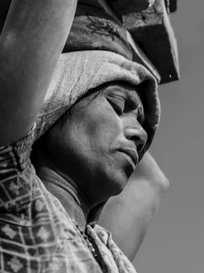 A close-up monochrome portrait of a woman worker, her eyes closed in exertion. The photo captures the immense physical and mental toll of her daily labor.