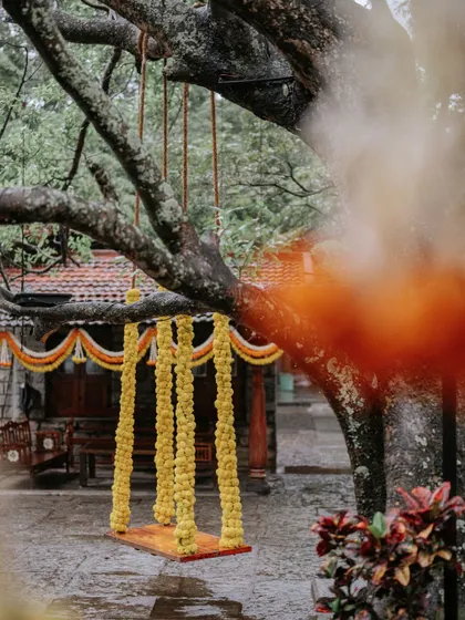A whimsical floral swing decorated with marigold garlands, set against the rustic backdrop of the venue. The orange smoke adds a touch of drama and color to the scene.