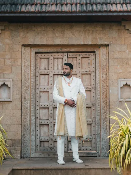 The groom stands tall and proud in his white sherwani, posing in front of an ornate wooden door, looking every bit the royal groom.
