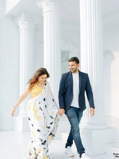 A happy, carefree moment during a pre-wedding shoot. The couple walks hand-in-hand through a beautiful white colonnade, their smiles lighting up the frame.