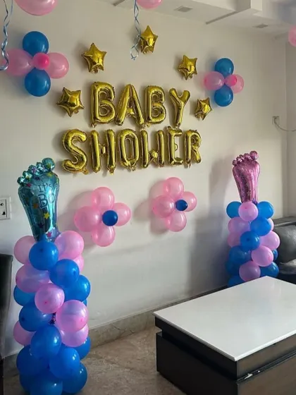 A baby shower decoration in a living room. The setup includes gold foil 'BABY SHOWER' balloons and pillars with foot-shaped foil balloons.