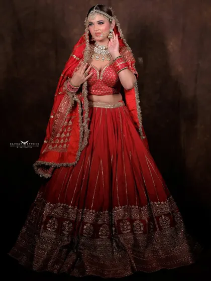 A full-length studio portrait of a bride in a classic red lehenga. The dark background makes the vibrant red and gold embroidery pop.