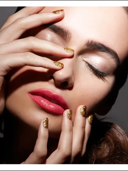 A close-up beauty shot focusing on the contrast between bold red lips and sparkling gold nails. The pose is soft and intimate, drawing attention to the color and texture.