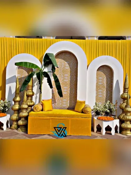 The main seating area for a Haldi function, featuring a custom-made yellow sofa set against a backdrop of modern arches and traditional brass elements. The banana leaves add a touch of tropical tradition.