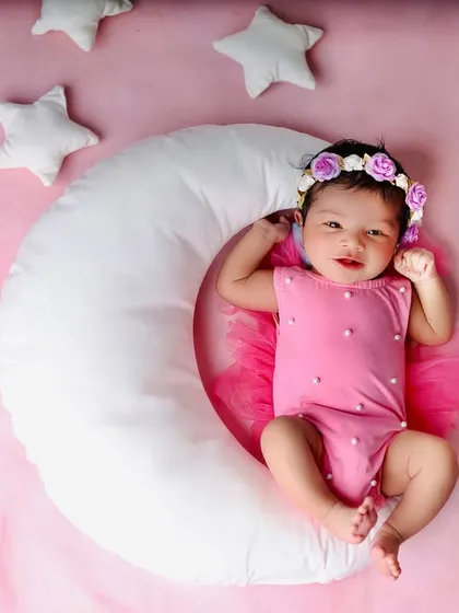 Over the moon for this little one. A sweet baby girl in a pink outfit rests on a soft crescent moon prop, surrounded by plush white stars for a dreamy, celestial-themed shot.