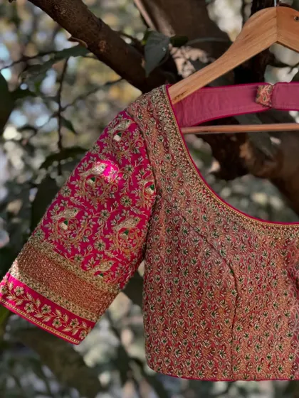 A close-up of a pink blouse featuring peacock motifs embroidered with silk thread and zardosi. The sleeve cuff has a thick, ornate border, adding to its grandeur.