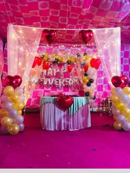 A classic anniversary stage setup under a tent. The decoration includes a 'Happy Anniversary' foil banner, heart-shaped balloons, and balloon columns in red, yellow, and white.