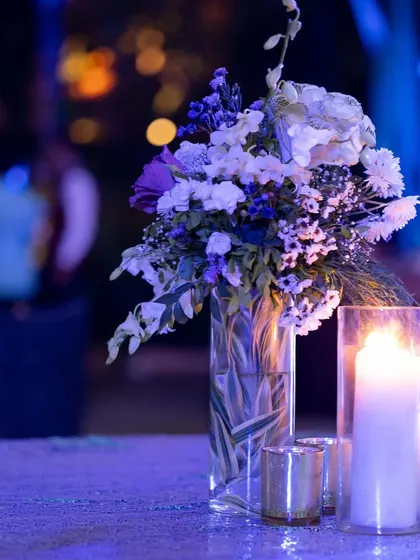 A beautiful centerpiece for a celestial-themed sangeet. A bouquet of white and purple flowers in a glass vase, surrounded by candles, creates a soft and romantic glow on the tables.