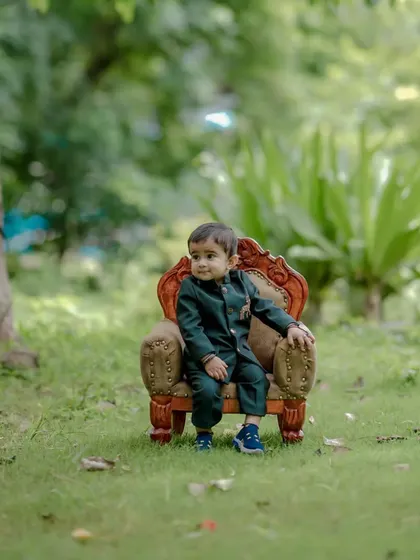 A wide shot showing the little boy in his smart suit, seated on the miniature throne in a beautiful park. This type of portrait captures both the child and the lovely outdoor environment of the shoot.