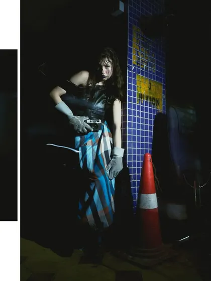 A full-body shot in a dark, industrial-looking setting, with the model holding a camera. The use of shadow and the gritty environment contribute to the cinematic, mysterious narrative of the series.