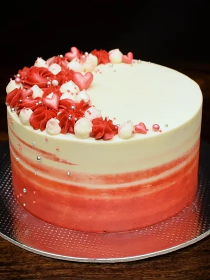 A beautiful red velvet anniversary cake with a red ombre watercolor effect. The top is decorated with a mix of red and white piped swirls and delicate heart-shaped sprinkles.