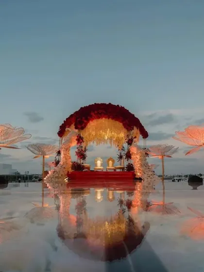 A dreamy mandap that appears to float on water. The reflection on the mirrored floor, combined with the red and white floral dome, creates an ethereal and magical setting for an evening ceremony.