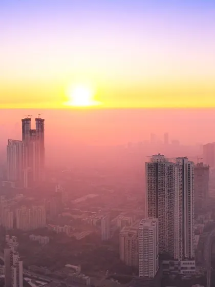 The sun appears as a bright orb in a hazy pink sky, setting behind the tall buildings of Mumbai.