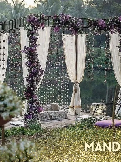 An example of a beautiful mandap we can create, with purple flowers and white drapes set against a serene natural backdrop.