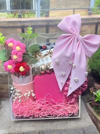 This vibrant pink gift set is presented in a clear acrylic box, showcasing all the beautiful items inside. It's filled with pink shredded paper and includes a variety of accessories and treats.
