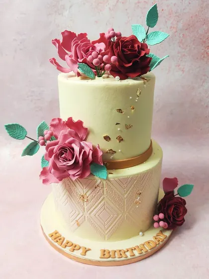 We showcase our artistry with this two-tiered cake featuring handmade edible sugar flowers. Intricate red and pink roses with delicate teal leaves adorn the cake, which also features an elegant embossed pattern on the bottom tier, perfect for a wedding or engagement.