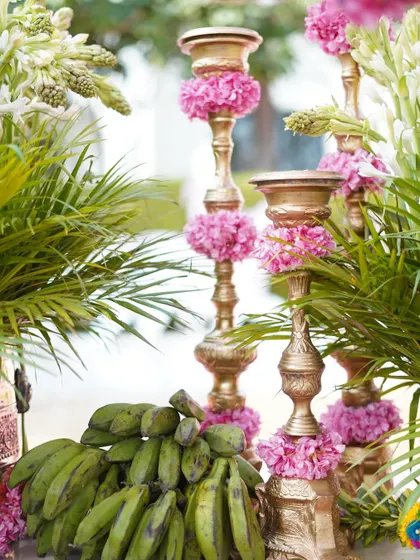 A unique detail combining rustic and traditional elements. Golden candle stands are adorned with pink flowers and placed next to bunches of green bananas, a symbol of prosperity.