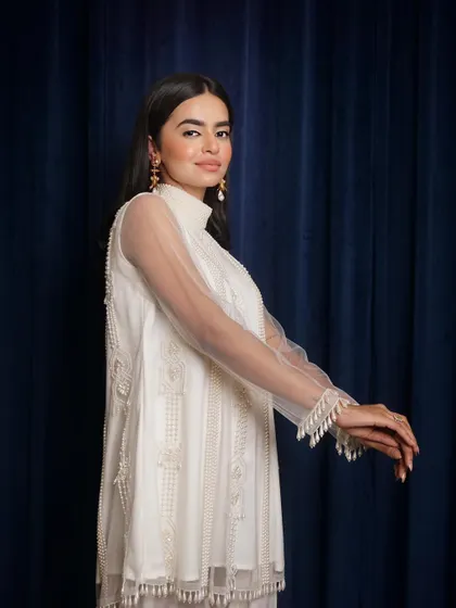 An elegant profile shot of a model in a white tunic with sheer, detailed sleeves. The deep blue curtain provides a rich, contrasting background.