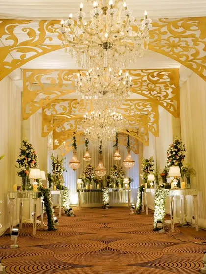 A grand, golden-themed entrance corridor for a luxurious reception. The design features intricate gold laser-cut panels, tall floral arrangements, and warm lighting to create an opulent atmosphere.