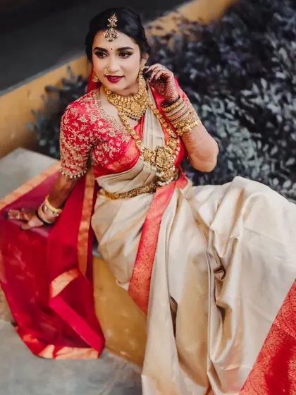 A beautiful portrait of the bride seated, showcasing her stunning red and cream saree and the intricate details of her gold jewellery.