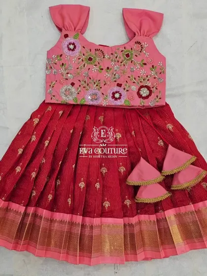 A sleeveless pattu pavadai for toddlers. The pink blouse has beautiful floral embroidery, and it's paired with a red pleated skirt.