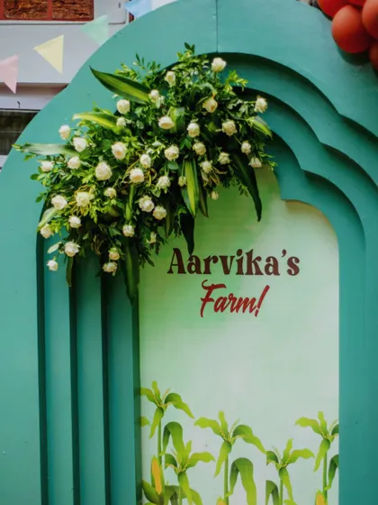 A close-up of the personalized "Aarvika's Farm" sign, framed by a beautiful teal arch and a spray of white roses. The design combines rustic charm with elegant touches.