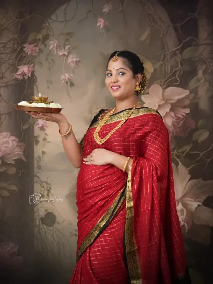 A divine studio portrait of a mother-to-be in a traditional red saree, holding a plate of offerings (puja thali).