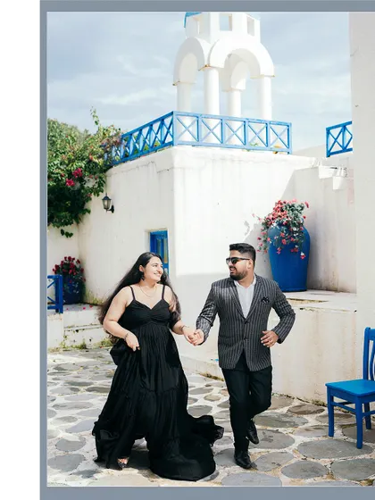 A playful pre-wedding shot of a couple running through a Santorini-themed set, their black outfits contrasting beautifully with the white and blue architecture.