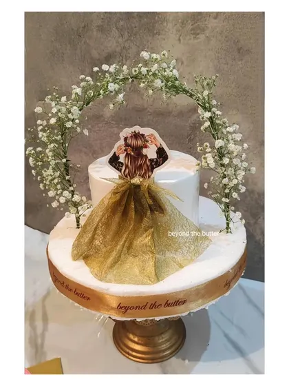 A dreamy "Bride to be" cake featuring a beautiful floral arch made of baby's breath. The cake is finished with a custom topper showing the bride in a flowing golden dress.