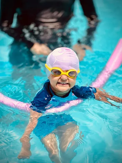 Using a pool noodle for support, this young swimmer is learning to float and balance with a big grin. Flotation aids are excellent tools for beginners.