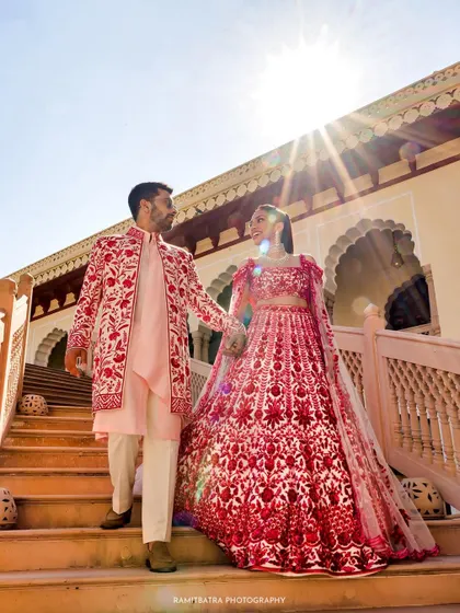 A couple in coordinated red and pink Manish Malhotra outfits descends the grand staircase at Rambagh Palace, a perfect blend of modern style and royal heritage.