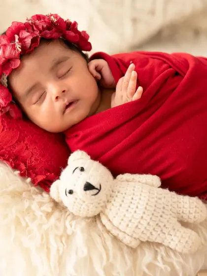 A sweet close-up of the newborn girl in her red wrap, cuddling with a small white teddy bear.